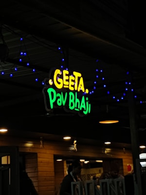 A restaurant sign reads 'Geeta Pav Bhaji' in bright yellow and green letters. Blue decorative lights are strung around the sign, adding a festive appearance. The background shows a wooden exterior with warm lighting overhead, suggesting an inviting atmosphere. People are partially visible, indicating activity inside the establishment.