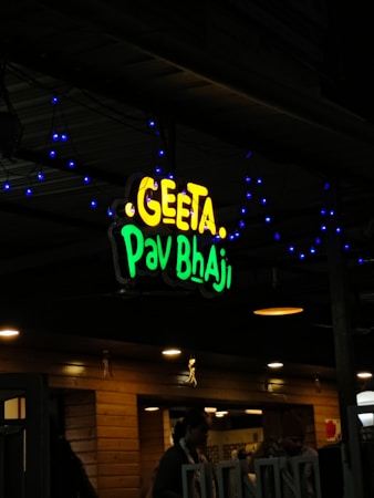 A restaurant sign reads 'Geeta Pav Bhaji' in bright yellow and green letters. Blue decorative lights are strung around the sign, adding a festive appearance. The background shows a wooden exterior with warm lighting overhead, suggesting an inviting atmosphere. People are partially visible, indicating activity inside the establishment.