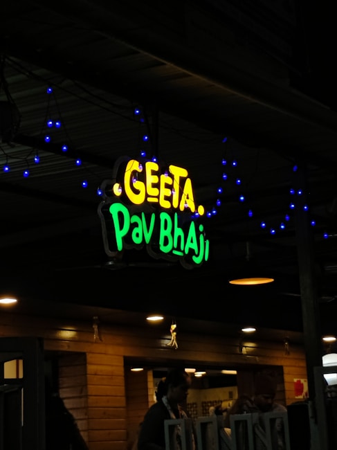A restaurant sign reads 'Geeta Pav Bhaji' in bright yellow and green letters. Blue decorative lights are strung around the sign, adding a festive appearance. The background shows a wooden exterior with warm lighting overhead, suggesting an inviting atmosphere. People are partially visible, indicating activity inside the establishment.