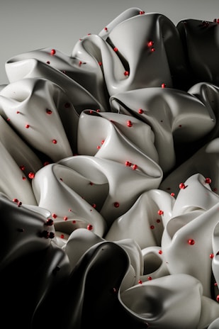 A close-up view of folded, silk-like fabric with a smooth, matte texture, scattered with small, glossy red spheres. The folds create an undulating, abstract pattern.