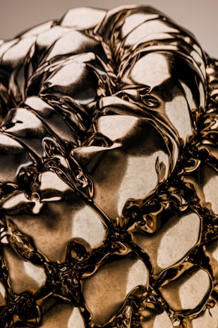 A close-up of a highly reflective, metallic surface with intricate folds and creases, giving it a fluid-like appearance. The surface captures and reflects light, creating varying shades and highlights.