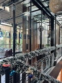 A modern, stylish interior space featuring glass walls and wooden accents. The area is decorated with festive garlands adorned with ornaments and lights. There are plants visible outside the glass, adding a touch of greenery. Warm and cozy lighting enhances the inviting atmosphere.