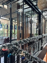 A modern, stylish interior space featuring glass walls and wooden accents. The area is decorated with festive garlands adorned with ornaments and lights. There are plants visible outside the glass, adding a touch of greenery. Warm and cozy lighting enhances the inviting atmosphere.