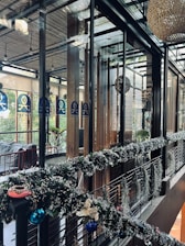 A modern, stylish interior space featuring glass walls and wooden accents. The area is decorated with festive garlands adorned with ornaments and lights. There are plants visible outside the glass, adding a touch of greenery. Warm and cozy lighting enhances the inviting atmosphere.