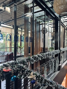 A modern, stylish interior space featuring glass walls and wooden accents. The area is decorated with festive garlands adorned with ornaments and lights. There are plants visible outside the glass, adding a touch of greenery. Warm and cozy lighting enhances the inviting atmosphere.