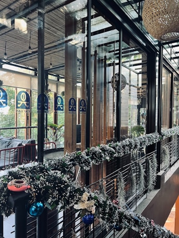 A modern, stylish interior space featuring glass walls and wooden accents. The area is decorated with festive garlands adorned with ornaments and lights. There are plants visible outside the glass, adding a touch of greenery. Warm and cozy lighting enhances the inviting atmosphere.