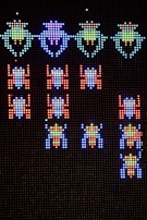 A pixelated display features rows of colorful, retro-style alien creatures from a classic arcade game. The aliens are arranged in formation against a dark background, with vibrant colors that include blue, green, red, and purple. The design is reminiscent of vintage gaming graphics, with a focus on simplicity and bold color contrasts.