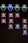 A pixelated display features rows of colorful, retro-style alien creatures from a classic arcade game. The aliens are arranged in formation against a dark background, with vibrant colors that include blue, green, red, and purple. The design is reminiscent of vintage gaming graphics, with a focus on simplicity and bold color contrasts.
