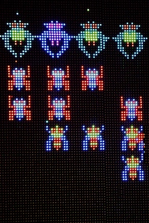 A pixelated display features rows of colorful, retro-style alien creatures from a classic arcade game. The aliens are arranged in formation against a dark background, with vibrant colors that include blue, green, red, and purple. The design is reminiscent of vintage gaming graphics, with a focus on simplicity and bold color contrasts.