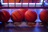 Several basketballs are aligned in a row on a basketball arcade machine platform. The environment has a colorful and illuminated backdrop with a mix of red and blue lighting.