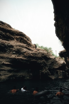 A group of people are swimming in a dark, natural body of water inside a rocky cave. The entrance of the cave is visible, allowing light to illuminate the rocky formations and creating a contrast between the dark water and the bright outdoors. Palm trees can be seen in the distance beyond the cave entrance. One person stands on a ledge, possibly preparing to jump into the water.