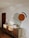 A modern room showcases a minimalist decor with a wooden sideboard against a white wall. The wall features abstract geometric art including a large green semi-circle and a smaller orange circle, accompanied by a black arc. On top of the sideboard are books arranged upright, decorative vases, and tall, slender candlesticks. The natural light creates soft, wavy shadows on the wall.