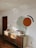 A modern room showcases a minimalist decor with a wooden sideboard against a white wall. The wall features abstract geometric art including a large green semi-circle and a smaller orange circle, accompanied by a black arc. On top of the sideboard are books arranged upright, decorative vases, and tall, slender candlesticks. The natural light creates soft, wavy shadows on the wall.