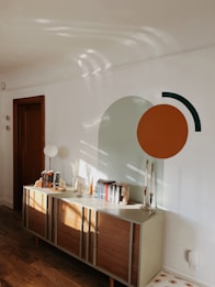 A modern room showcases a minimalist decor with a wooden sideboard against a white wall. The wall features abstract geometric art including a large green semi-circle and a smaller orange circle, accompanied by a black arc. On top of the sideboard are books arranged upright, decorative vases, and tall, slender candlesticks. The natural light creates soft, wavy shadows on the wall.
