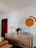 A modern living room with a wooden dresser against a white wall, featuring decorative items such as vases, candles, and a floral arrangement. On the wall, a large circular and semi-circular design is painted in orange and dark green. A closed wooden door and part of a tan leather sofa are visible.