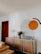 A modern living room with a wooden dresser against a white wall, featuring decorative items such as vases, candles, and a floral arrangement. On the wall, a large circular and semi-circular design is painted in orange and dark green. A closed wooden door and part of a tan leather sofa are visible.
