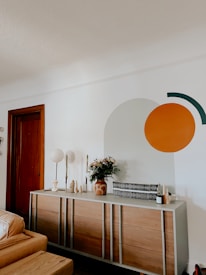 A modern living room with a wooden dresser against a white wall, featuring decorative items such as vases, candles, and a floral arrangement. On the wall, a large circular and semi-circular design is painted in orange and dark green. A closed wooden door and part of a tan leather sofa are visible.