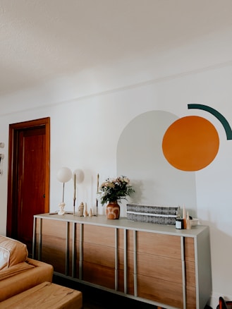 A modern living room with a wooden dresser against a white wall, featuring decorative items such as vases, candles, and a floral arrangement. On the wall, a large circular and semi-circular design is painted in orange and dark green. A closed wooden door and part of a tan leather sofa are visible.