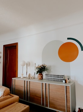 A modern living room with a wooden dresser against a white wall, featuring decorative items such as vases, candles, and a floral arrangement. On the wall, a large circular and semi-circular design is painted in orange and dark green. A closed wooden door and part of a tan leather sofa are visible.