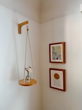 In a corner decorated with a small hanging shelf suspended by ropes, a vase adorned with a couple of flowers sits. Two framed artworks are hung on the adjacent wall; one features a pattern of green and yellow leaves while the other depicts a circular design with lines extending below it.