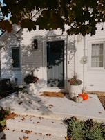 A porch with white siding features a dark green door and two planters filled with autumn foliage. Two pumpkins, one orange and one white, sit on the ground near the door. Fallen leaves are scattered across the porch and steps, with shadows from overhanging tree branches creating a dappled effect.