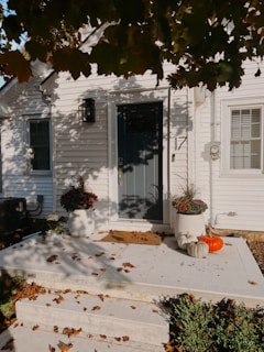A porch with white siding features a dark green door and two planters filled with autumn foliage. Two pumpkins, one orange and one white, sit on the ground near the door. Fallen leaves are scattered across the porch and steps, with shadows from overhanging tree branches creating a dappled effect.