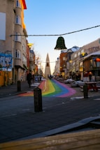 A vibrant urban street scene with a rainbow-painted pathway leading towards a large, striking church in the distance. Surrounding buildings are adorned with colorful murals and decorated with festive lights. The atmosphere is lively with the pastel shades of the buildings contrasting against a clear blue sky.