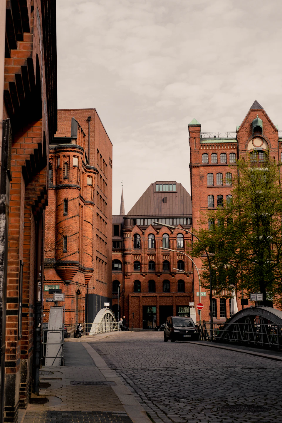 The image depicts a cobblestone street lined with brick buildings, including an old red brick building on the left side and another large brick building on the right side. The sky above is overcast, casting a soft light over the scene. A black car is parked on the right side of the street, and two people can be seen walking on the sidewalk in front of one of the buildings.