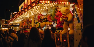 A bustling fairground booth is illuminated with bright, colorful lights. Large stuffed animals like bears are prominently displayed as prizes. A crowd of people, mostly seen from behind, gathers around the booth, creating a lively and festive atmosphere.