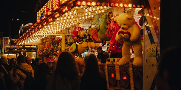 A bustling fairground booth is illuminated with bright, colorful lights. Large stuffed animals like bears are prominently displayed as prizes. A crowd of people, mostly seen from behind, gathers around the booth, creating a lively and festive atmosphere.