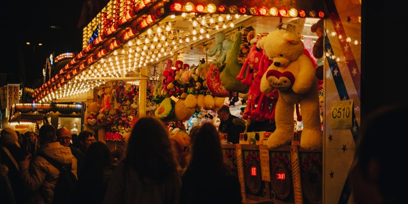 A bustling fairground booth is illuminated with bright, colorful lights. Large stuffed animals like bears are prominently displayed as prizes. A crowd of people, mostly seen from behind, gathers around the booth, creating a lively and festive atmosphere.