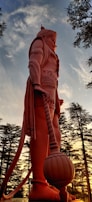A towering statue of a mythological character, depicted in a heroic stance with intricate armor and a large mace. The background features a serene sunset with a sky of soft blues and warm oranges, framed by tall, slender trees.