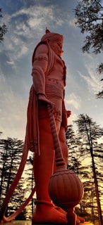 A towering statue of a mythological character, depicted in a heroic stance with intricate armor and a large mace. The background features a serene sunset with a sky of soft blues and warm oranges, framed by tall, slender trees.
