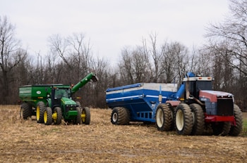 Two large farm tractors are parked in a recently harvested field. The tractor on the left is green with yellow wheels and attached to a green grain cart. The tractor on the right is red with off-white wheels, connected to a blue grain cart. Surrounding the tractors, the field is covered with dried corn stalks and bordered by bare trees against a cloudy sky.