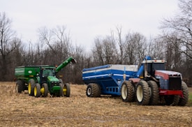 Two large farm tractors are parked in a recently harvested field. The tractor on the left is green with yellow wheels and attached to a green grain cart. The tractor on the right is red with off-white wheels, connected to a blue grain cart. Surrounding the tractors, the field is covered with dried corn stalks and bordered by bare trees against a cloudy sky.