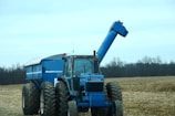A blue tractor with large tires is parked in a field with an empty trailer attached. The field has remnants of harvested crops, and there are leafless trees in the background under a cloudy sky.