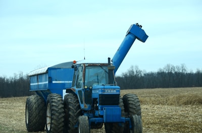 A blue tractor with large tires is parked in a field with an empty trailer attached. The field has remnants of harvested crops, and there are leafless trees in the background under a cloudy sky.