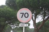 A round road sign displaying the number 70, possibly indicating a speed limit, set against a background of dense green pine trees. Below the sign, another smaller sign reads 'LEMBRE-SE,' which means 'remember' in Portuguese.