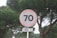 A round road sign displaying the number 70, possibly indicating a speed limit, set against a background of dense green pine trees. Below the sign, another smaller sign reads 'LEMBRE-SE,' which means 'remember' in Portuguese.