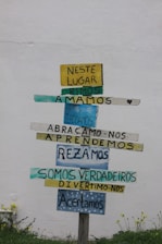 A wooden signpost against a plain white wall displays multiple colorful horizontal planks with Portuguese text. Each plank contains different words in varying colors like yellow, blue, and teal. The ground below has small green plants and yellow flowers.