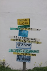 A wooden signpost against a plain white wall displays multiple colorful horizontal planks with Portuguese text. Each plank contains different words in varying colors like yellow, blue, and teal. The ground below has small green plants and yellow flowers.