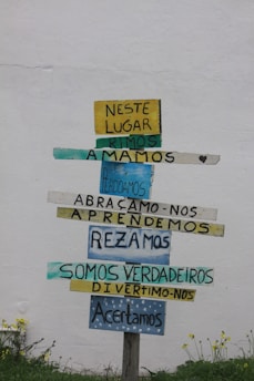 A wooden signpost against a plain white wall displays multiple colorful horizontal planks with Portuguese text. Each plank contains different words in varying colors like yellow, blue, and teal. The ground below has small green plants and yellow flowers.