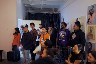 A group of people is gathered in an indoor space, possibly for an event or exhibition. They appear to be attentively listening or watching something. The walls are adorned with various artworks, suggesting that this could be an art gallery or studio. The setting is casual, and the people are dressed in a range of casual to semi-casual clothes. The background includes a black plastic curtain and some exposed pipes on the ceiling.