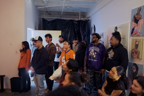 A group of people is gathered in an indoor space, possibly for an event or exhibition. They appear to be attentively listening or watching something. The walls are adorned with various artworks, suggesting that this could be an art gallery or studio. The setting is casual, and the people are dressed in a range of casual to semi-casual clothes. The background includes a black plastic curtain and some exposed pipes on the ceiling.