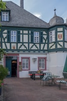 A traditional building with decorative timber framing and a slate roof, featuring small shuttered windows and intricate design elements. In front of the building, there is a courtyard with outdoor seating, including tables with chairs and a picnic bench, adorned with planters holding flowers.