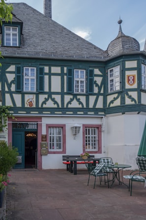 A traditional building with decorative timber framing and a slate roof, featuring small shuttered windows and intricate design elements. In front of the building, there is a courtyard with outdoor seating, including tables with chairs and a picnic bench, adorned with planters holding flowers.