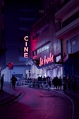 A nighttime scene featuring a cinema with neon lights. The marquee displays 'Los Ángeles,' and a crowd of people line up outside on the street. The area is illuminated by pink and purple lights, creating a vibrant atmosphere against the dark surroundings.
