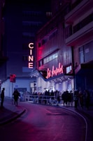 A nighttime scene featuring a cinema with neon lights. The marquee displays 'Los Ángeles,' and a crowd of people line up outside on the street. The area is illuminated by pink and purple lights, creating a vibrant atmosphere against the dark surroundings.
