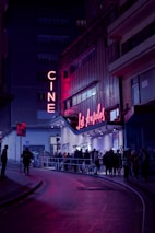 A nighttime scene featuring a cinema with neon lights. The marquee displays 'Los Ángeles,' and a crowd of people line up outside on the street. The area is illuminated by pink and purple lights, creating a vibrant atmosphere against the dark surroundings.
