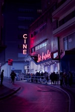 A nighttime scene featuring a cinema with neon lights. The marquee displays 'Los Ángeles,' and a crowd of people line up outside on the street. The area is illuminated by pink and purple lights, creating a vibrant atmosphere against the dark surroundings.
